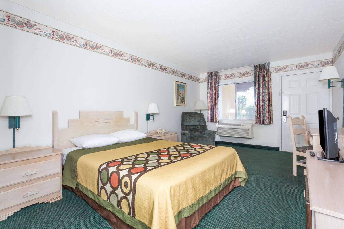 Una habitación de hotel con cama, una silla, un escritorio con computadora, televisor y ventana con cortinas.