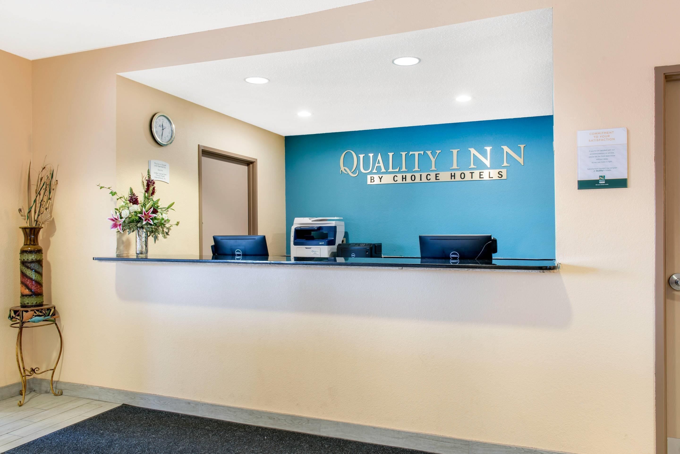 Un comptoir d’accueil avec une enseigne indiquant « Quality Inn by Choice Hotels », une horloge et un bureau équipé d’un ordinateur et d’une imprimante Dell.