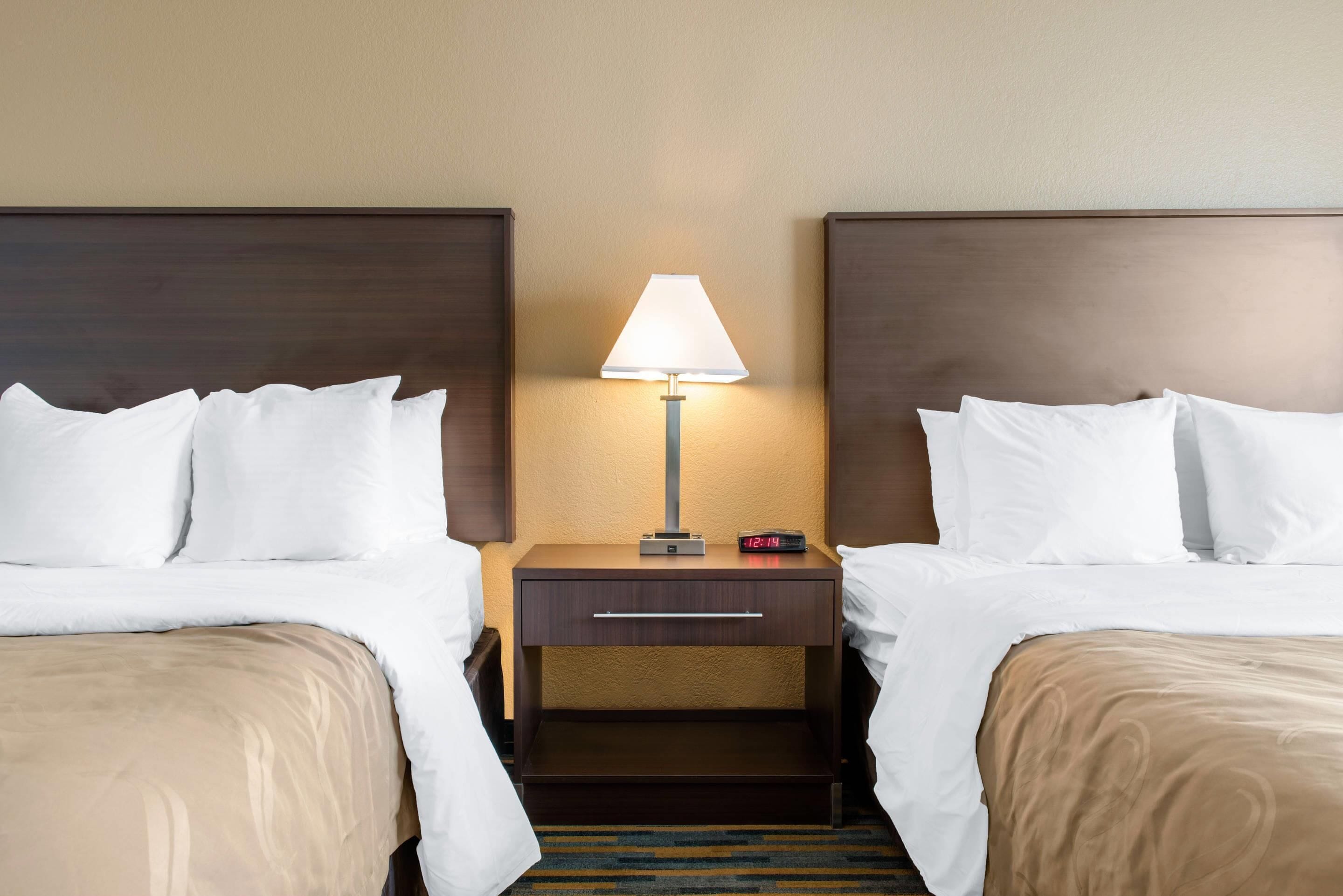 A hotel room with two beds, a nightstand with a lamp, and a digital clock.