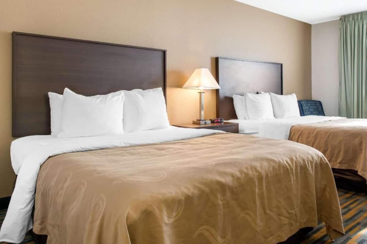 A hotel room with two beds, a nightstand with a lamp, and a view of the room.