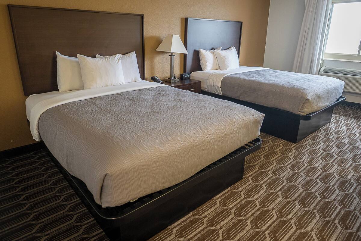 A hotel room with two beds, a nightstand with a lamp, and a window with curtains.