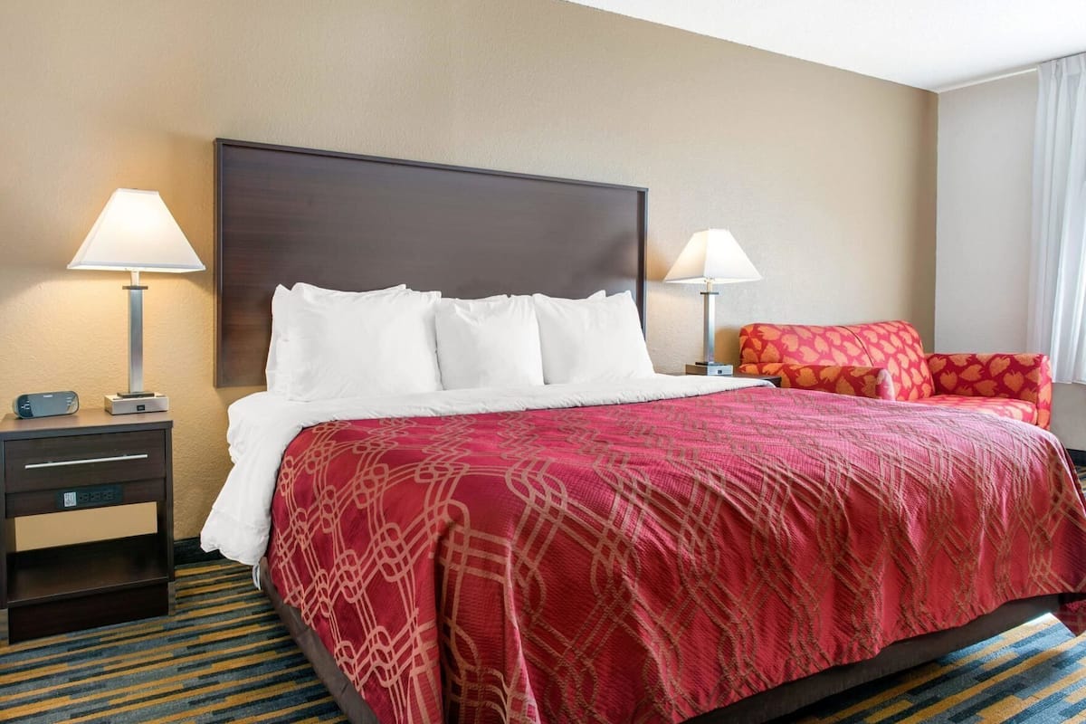 A hotel room with a large bed, two bedside tables, a nightstand, a red patterned bedspread, and a red and orange patterned sofa.
