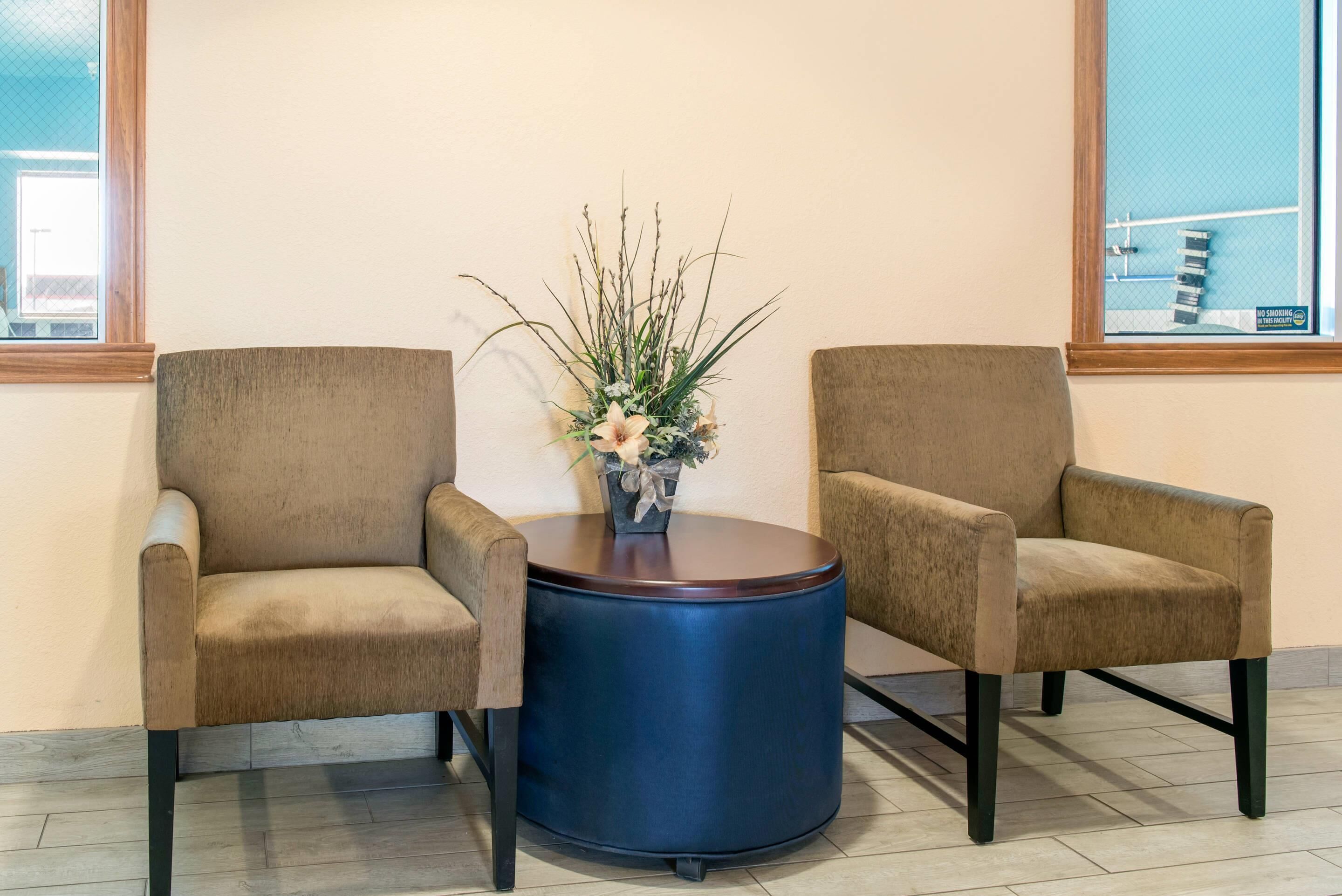 Deux fauteuils beiges sont placés face à face, séparés par une table bleu foncé. Sur la table, on aperçoit une petite plante en pot, et une fenêtre donne sur un parking situé à l’arrière-plan.