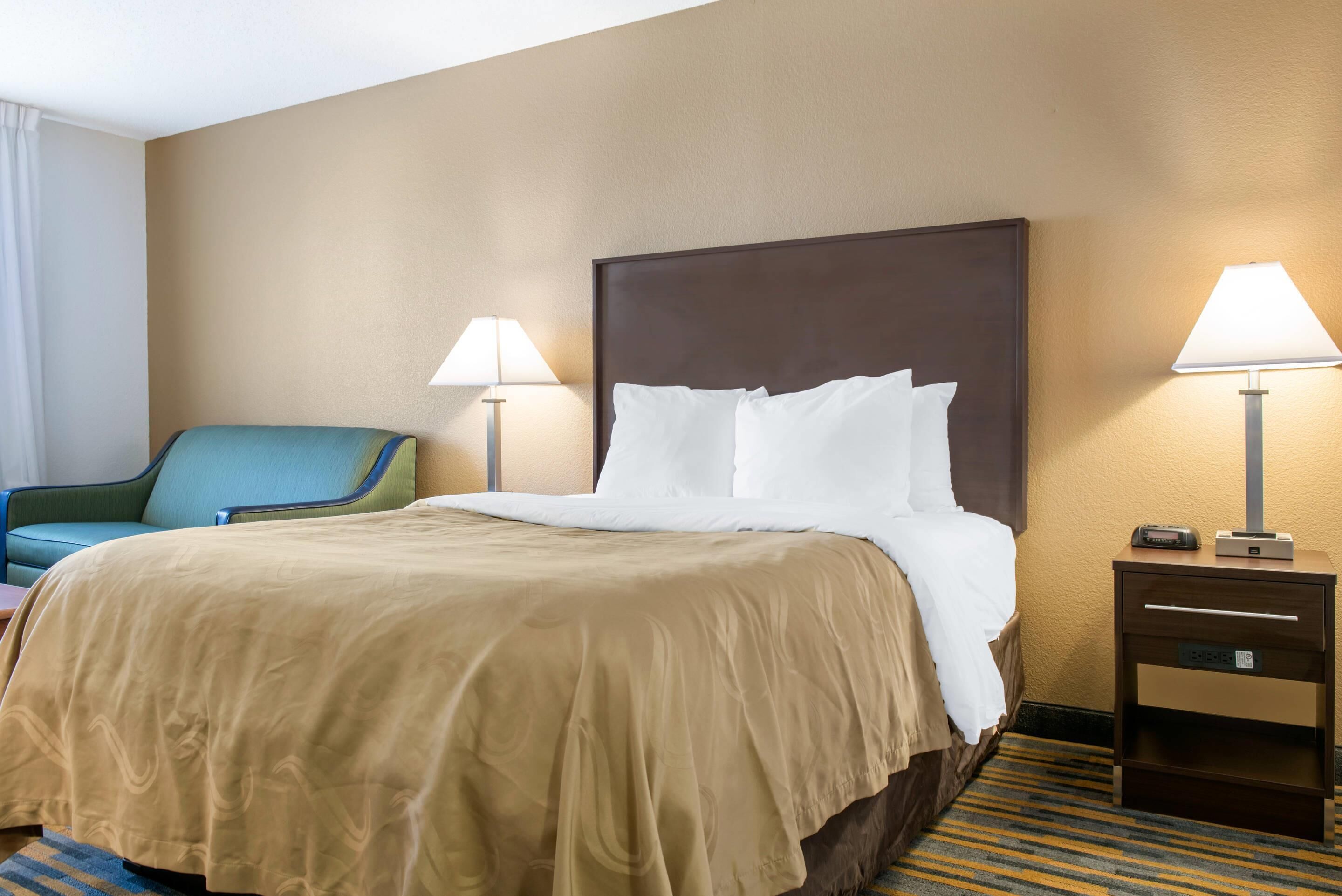 A hotel room with a large bed, two bedside lamps, a chair, a nightstand with a radio, and a carpeted floor.