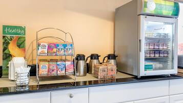 Un comptoir de cuisine où l’on trouve des boîtes de céréales, une machine à café et un réfrigérateur rempli de boissons.