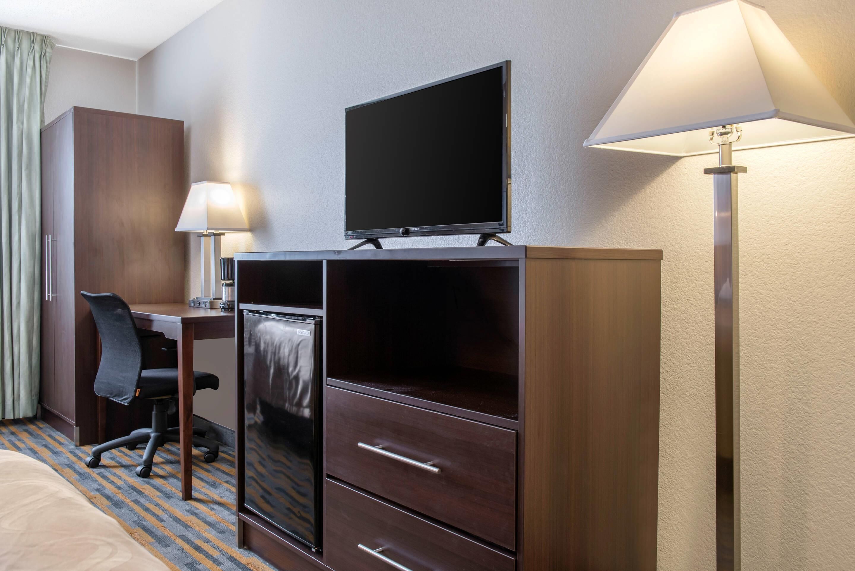 Una camera d'albergo con un letto, una scrivania con una sedia, una televisione, un minibar e una lampada.