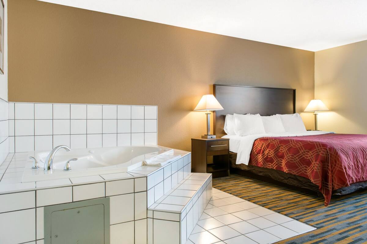 A hotel room with a large bathtub, two bedside lamps, a bed with a red blanket, and a patterned carpet.