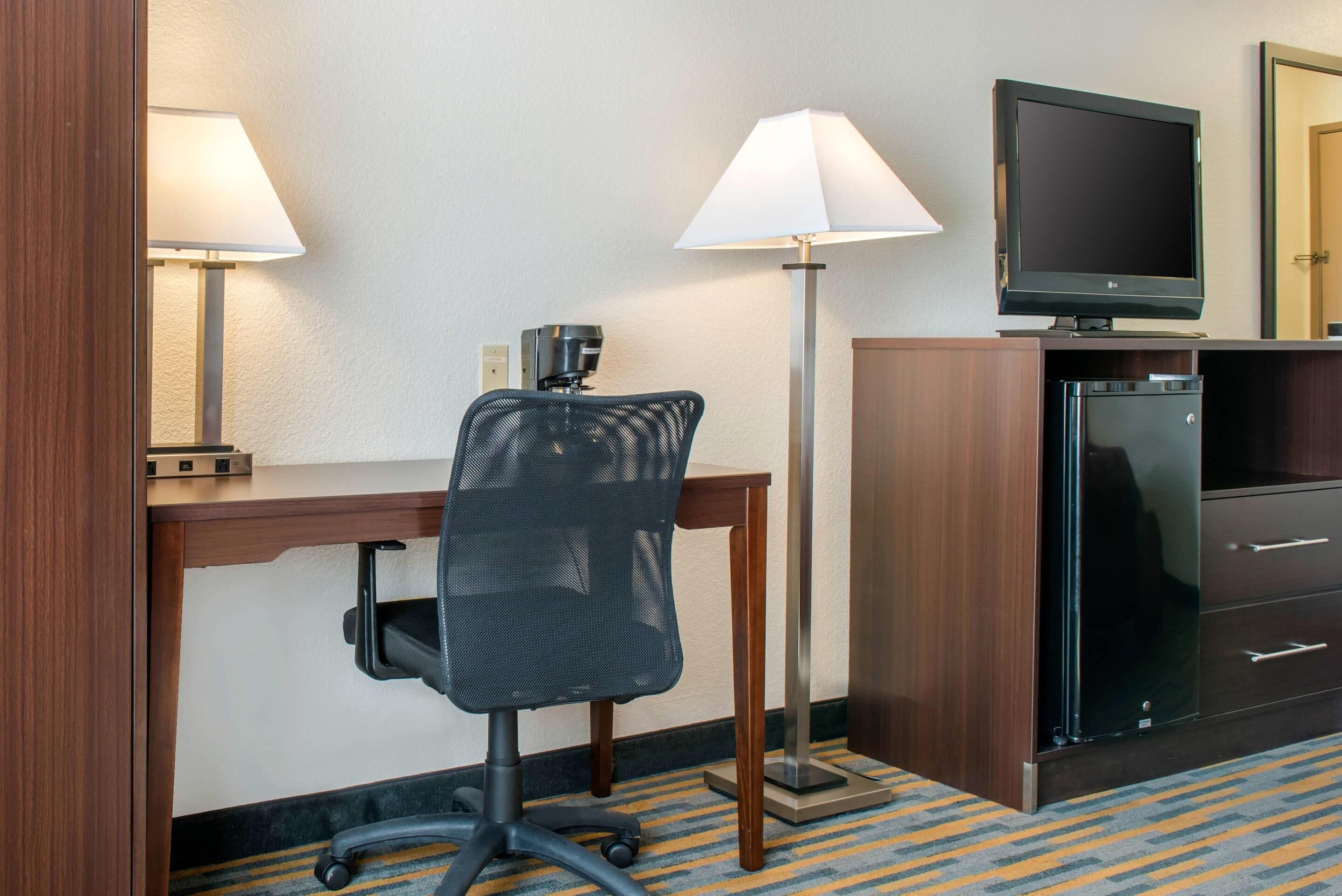 A hotel room with a desk, chair, television, and a minibar.