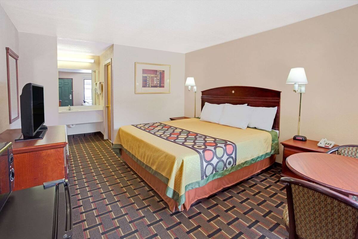 Habitación de hotel con cama, escritorio con televisor, una silla, lámpara y un cuadro en la pared.