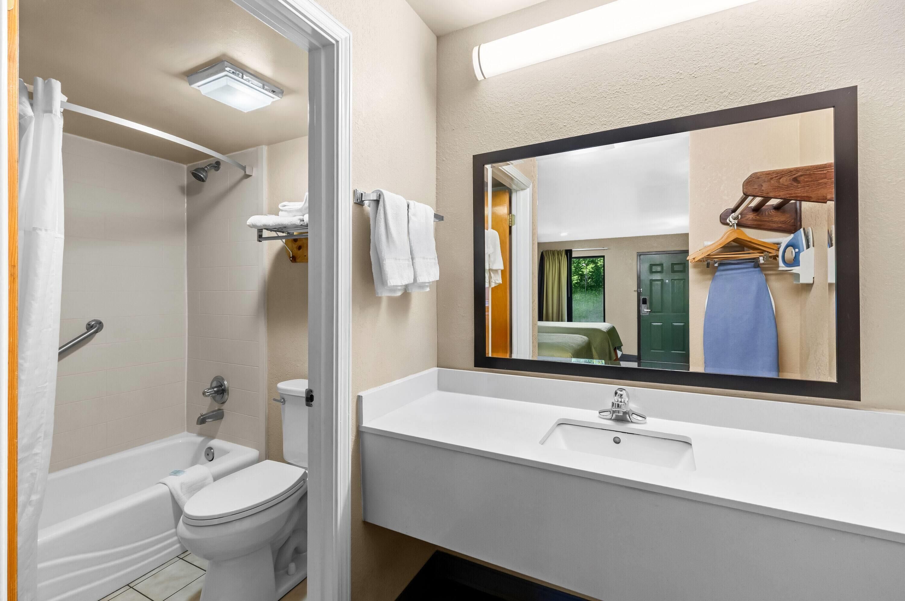 A bathroom with a bathtub, toilet, sink, and a mirror reflecting a room with a bed and a door.
