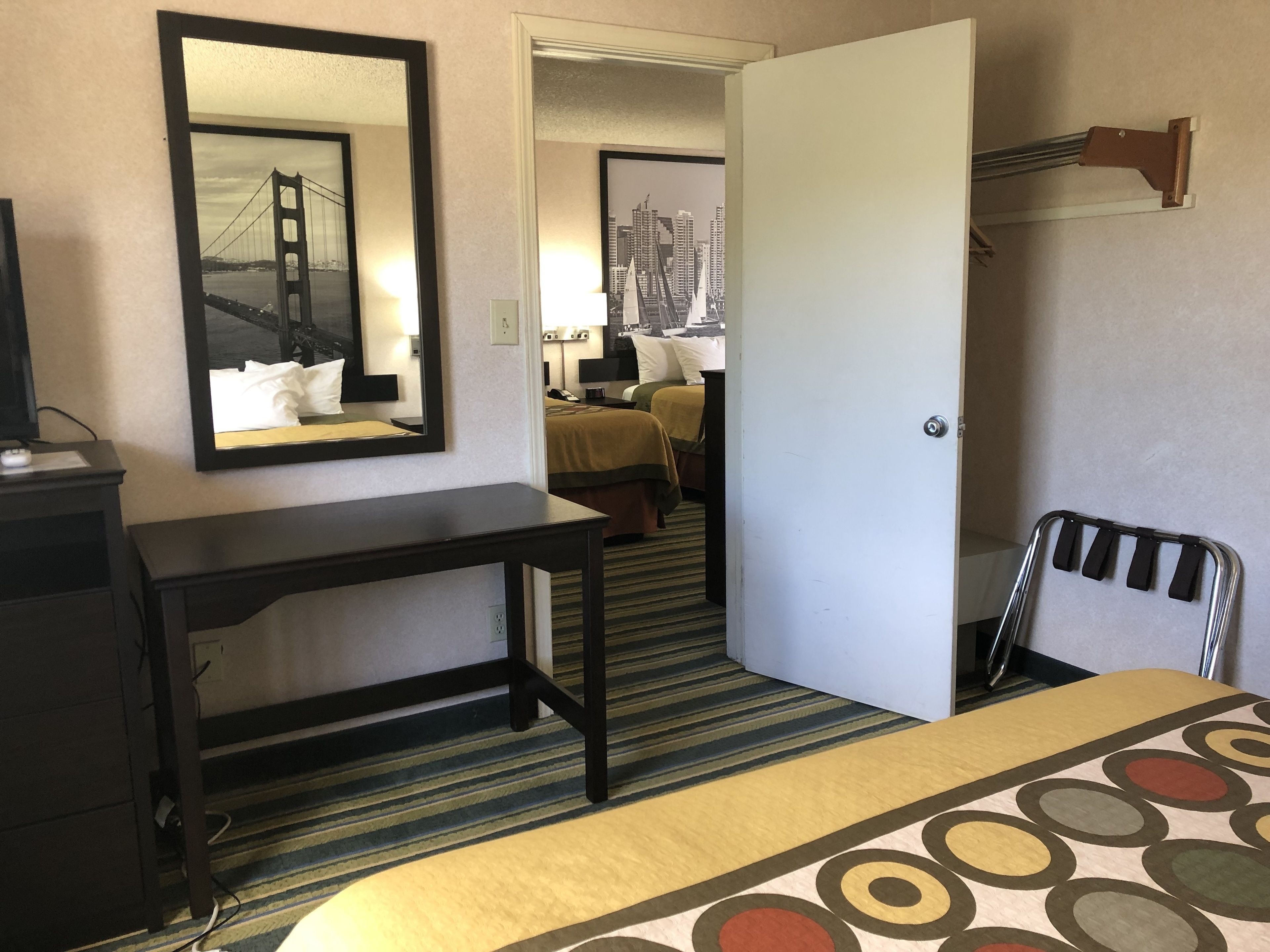 A hotel room with a bed, a desk, a chair, a mirror, and a door leading to another room.