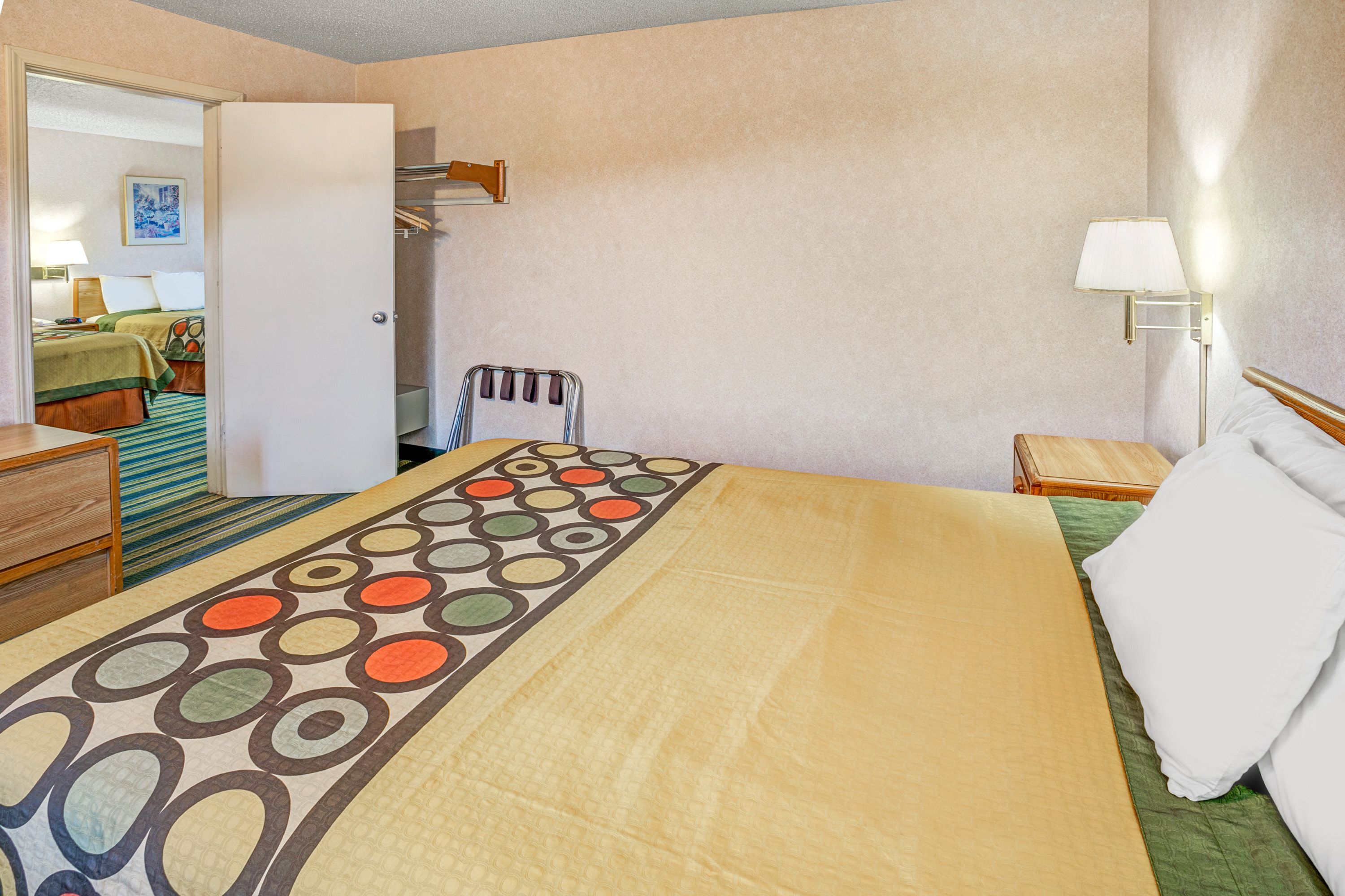 Habitación de hotel con una cama, una mesita de noche, una lámpara y vista a otra habitación con una cama y un cuadro en la pared.