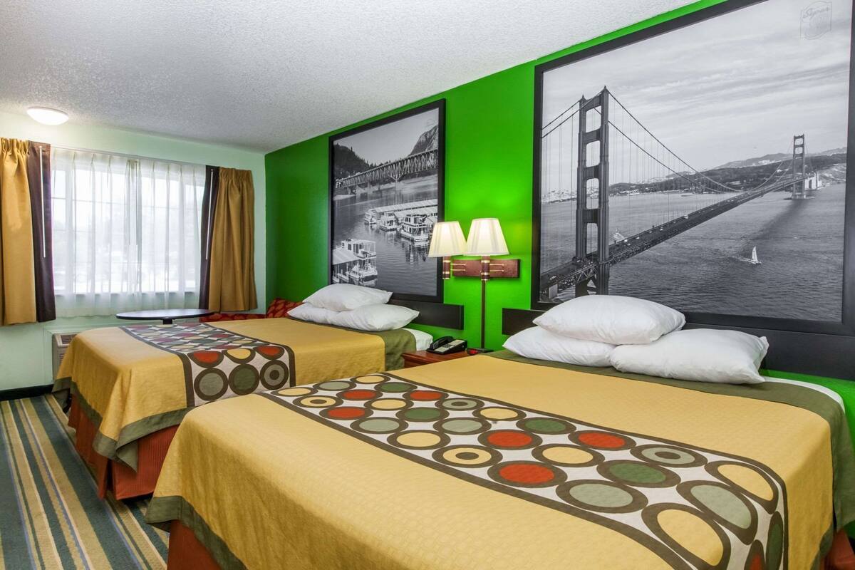 Habitació d'hotel amb dos llits, una finestra gran amb cortines, una paret d'accent verd i dos quadres grans amb imatges d'un pont.