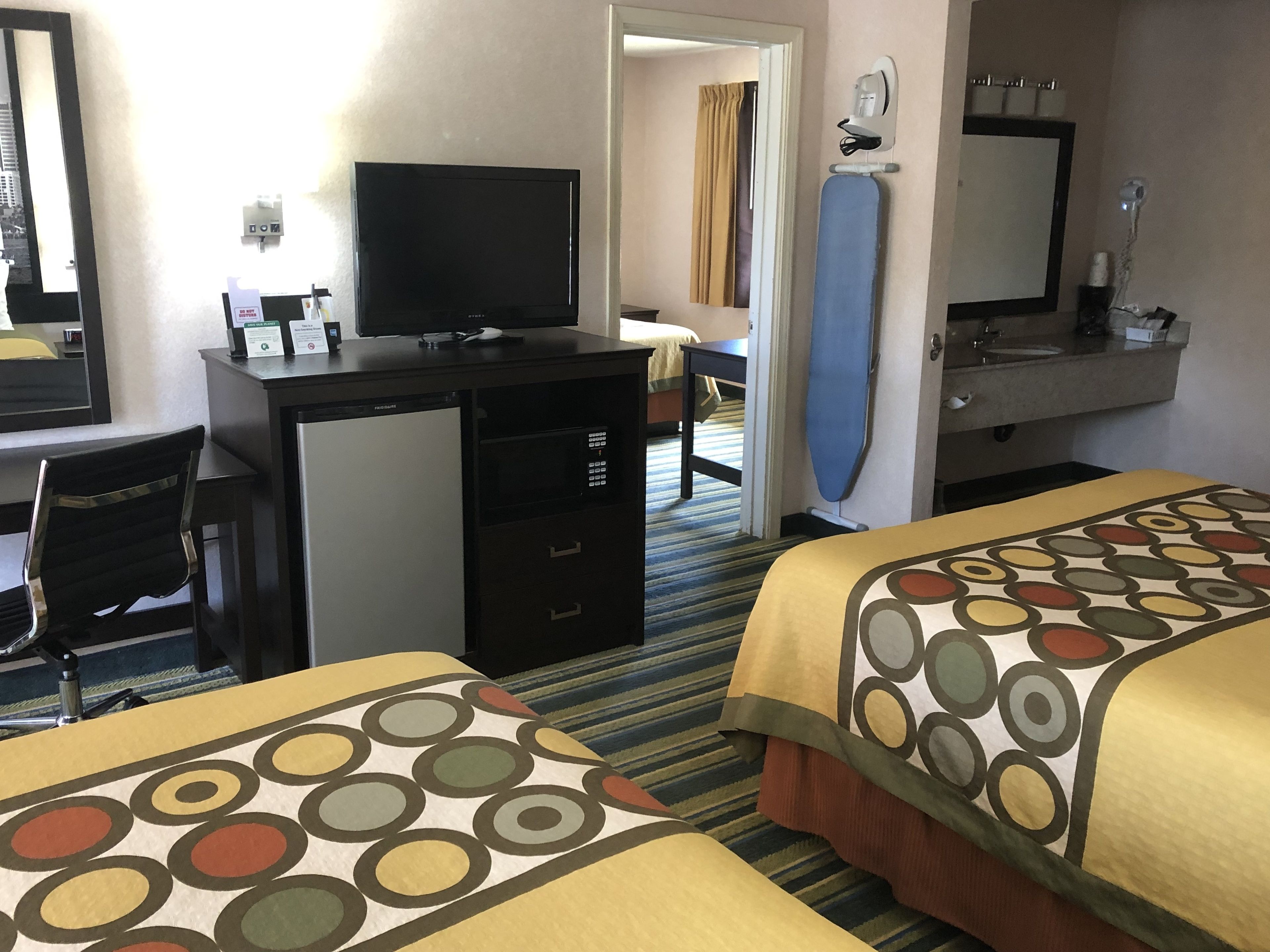 A hotel room with two beds, a desk with a computer, a small refrigerator, a mirror, and a bathroom with a sink and mirror.