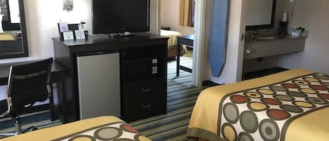 A hotel room with two beds, a desk with a computer, a small refrigerator, a mirror, and a bathroom with a sink and mirror.