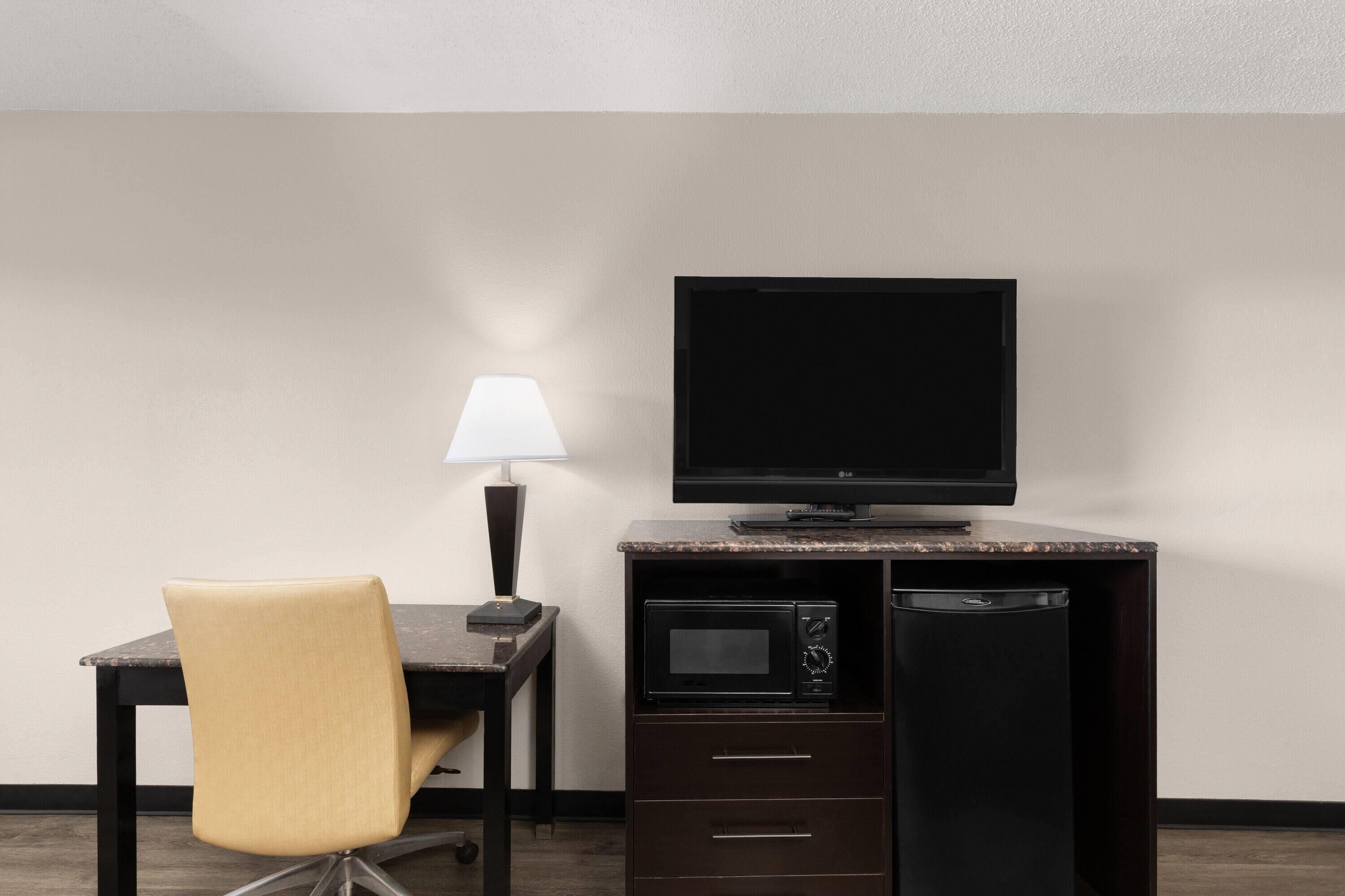 A compact hotel room with a desk, a chair, a flat-screen TV, a lamp, a microwave, and a refrigerator.