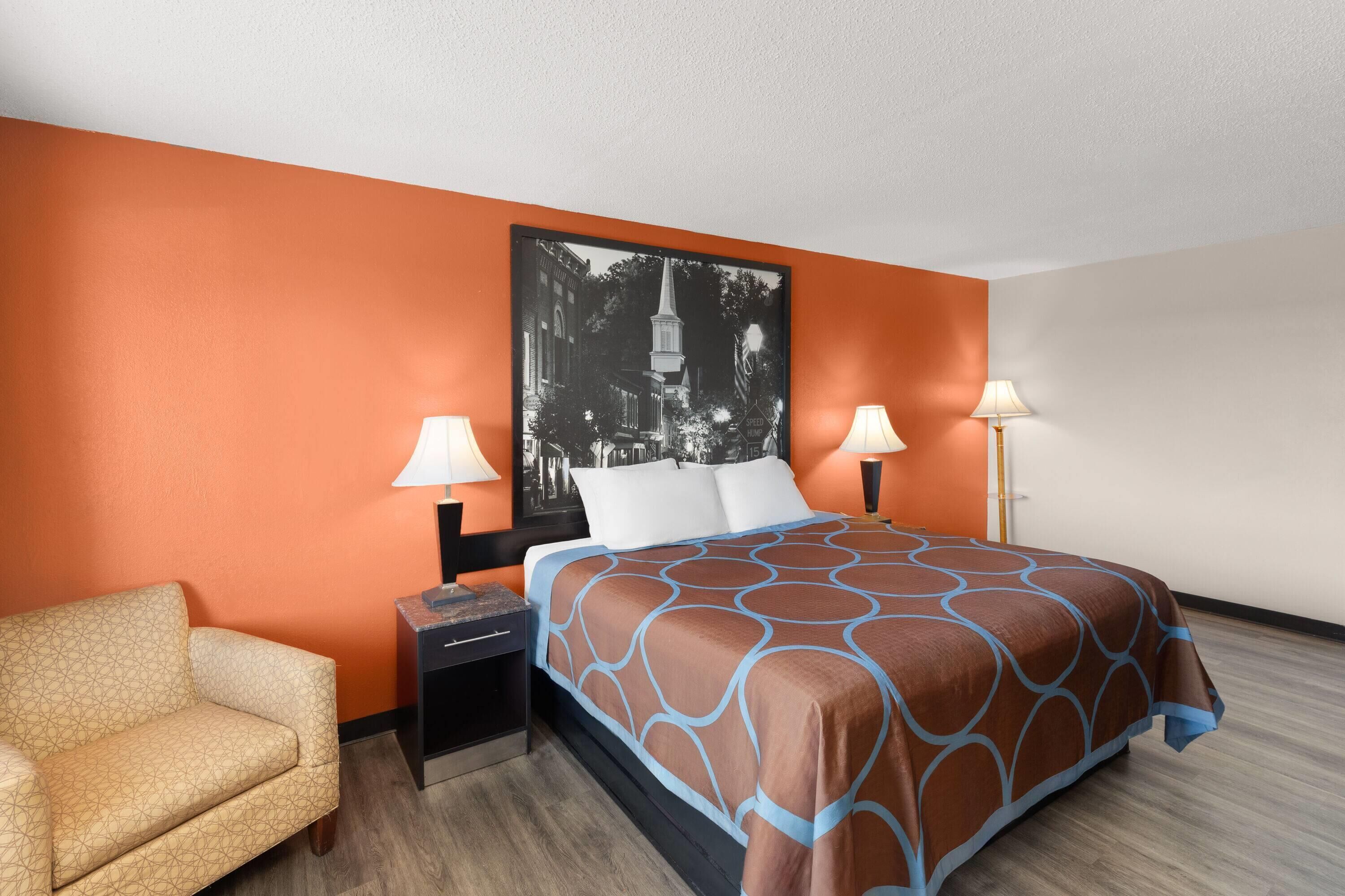 Een hotelkamer met een oranje wand, een bed met een beddeken met een patroon, twee bedlampen, een nachtkastje en een stoel.