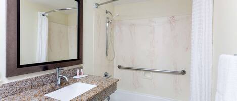 A bathroom with a granite countertop sink, a mirror, a shower with a grab bar, and a white bathtub.