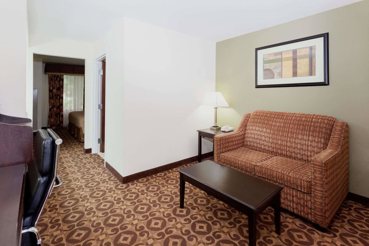 A hotel room with a patterned carpet, a sofa, a coffee table, a lamp, a framed picture on the wall, and a door leading to another room.