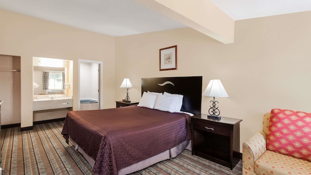 Habitación de hotel con una cama grande, una mesita de noche con lámparas, una silla con cojines estampados y una ventana con cortinas.