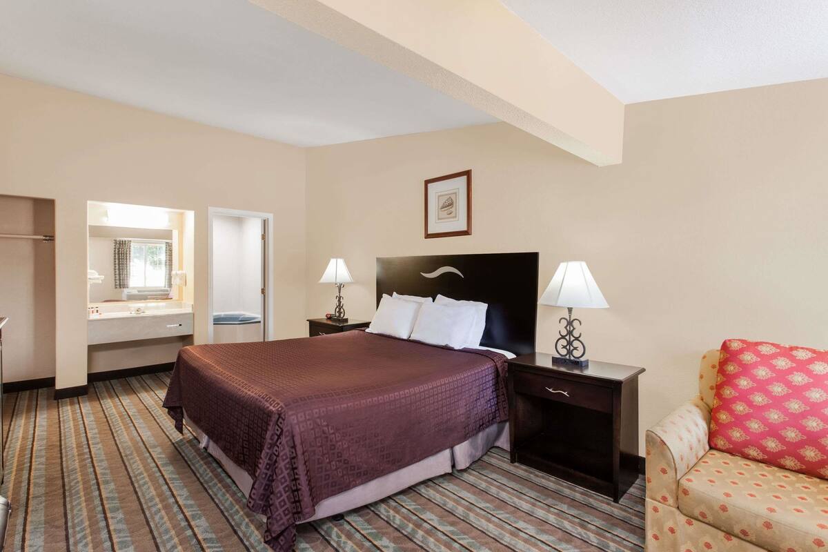 A hotel room with a large bed, a bedside table with lamps, a chair with patterned cushions, and a window with curtains.