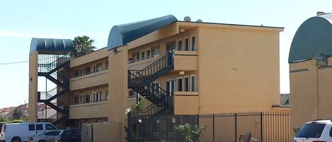 A multi-story building with a yellow facade, a blue roof, and a black staircase. There are cars parked in front and a fence along the side.