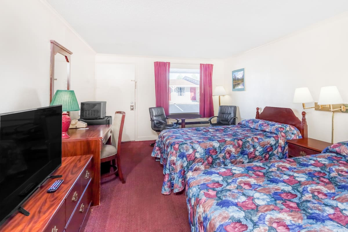 A hotel room with two beds, a TV, a desk with a lamp, and a window with curtains.