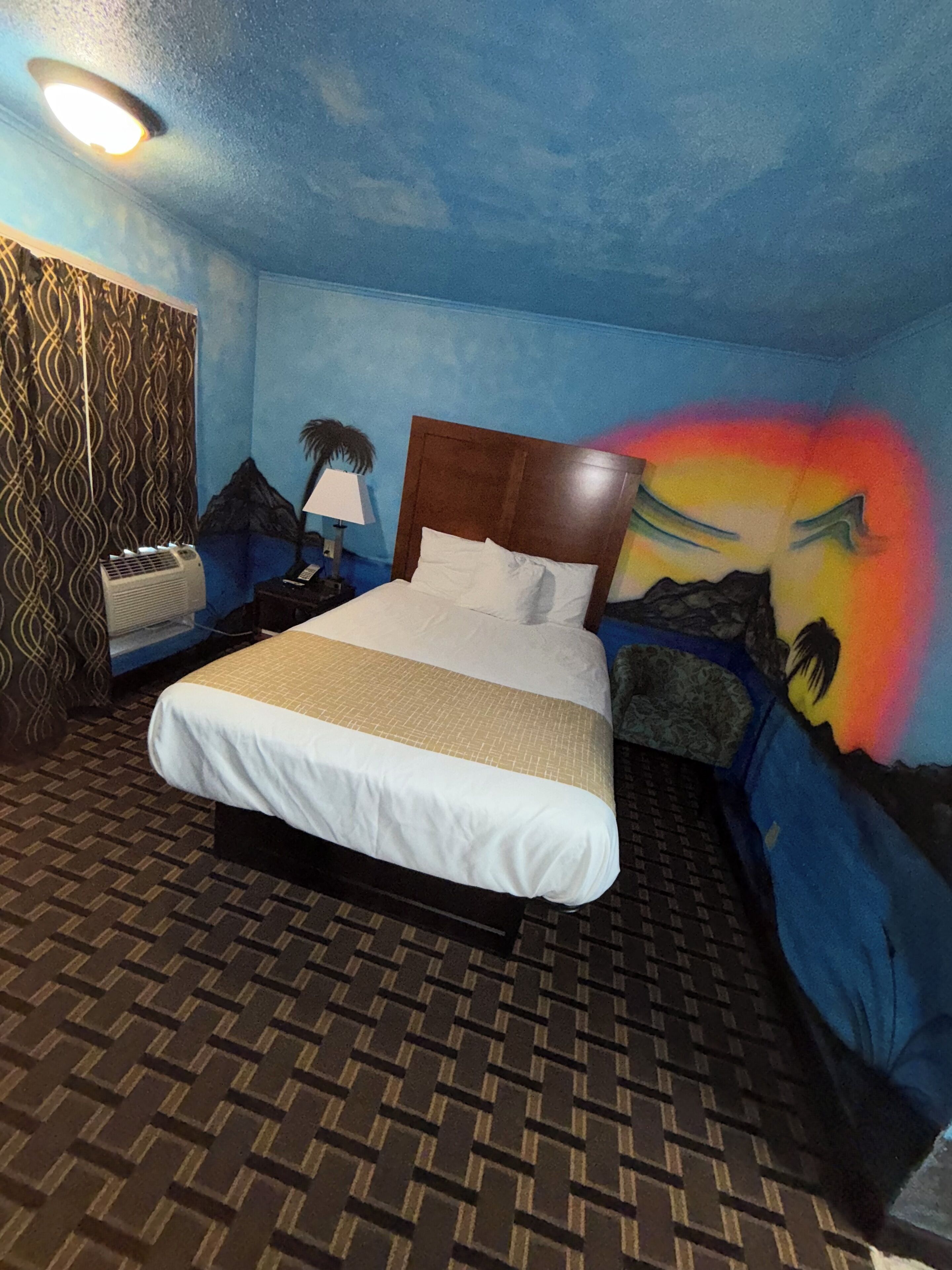 A hotel room with a bed, a nightstand, a lamp, a wall with a mountain mural, and a patterned carpet.