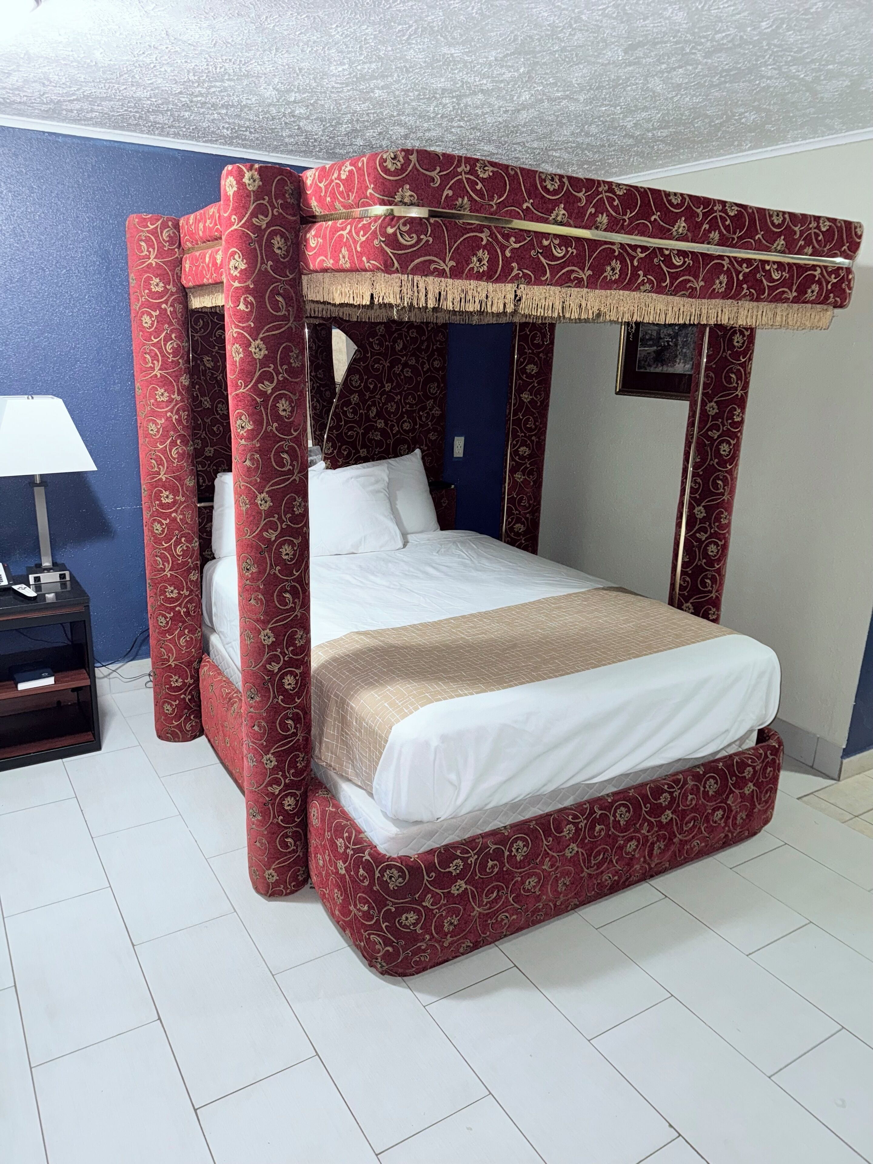 A four-poster bed with a red and gold patterned canopy, white bedding, and a bedside table with a lamp.
