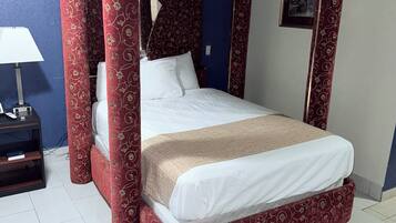 A four-poster bed with a red and gold patterned canopy, white bedding, and a bedside table with a lamp.