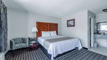 A hotel room with a bed, a chair, a nightstand, a lamp, a picture on the wall, and a bathroom visible through an open door.