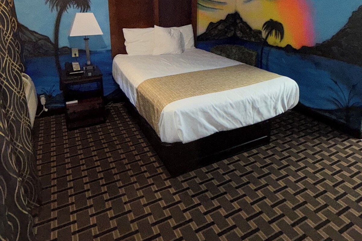 A hotel room with a bed, a nightstand, a lamp, and a mural of a sunset on the wall.