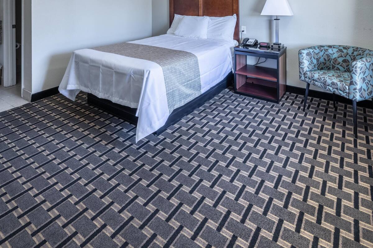 A hotel room with a bed, a nightstand, a lamp, a chair, and a patterned carpet.