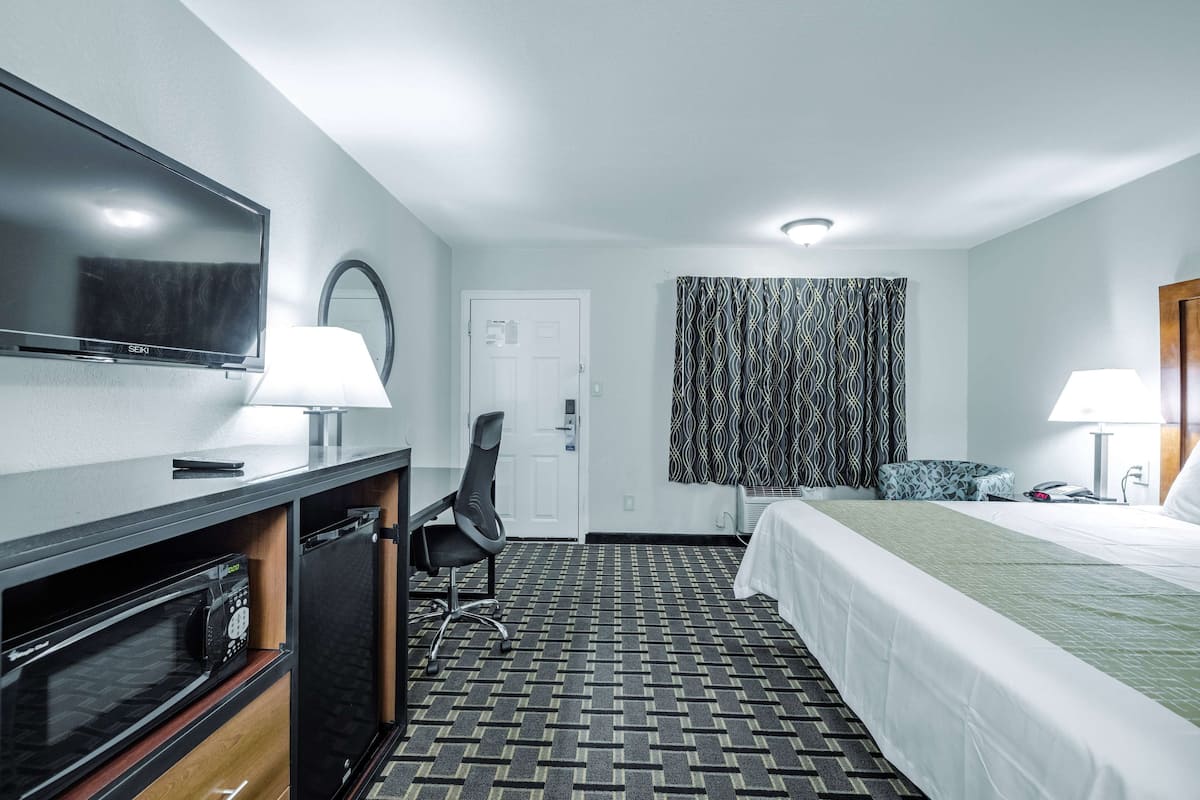 A hotel room with a bed, a desk with a television, a microwave, a chair, and a door.