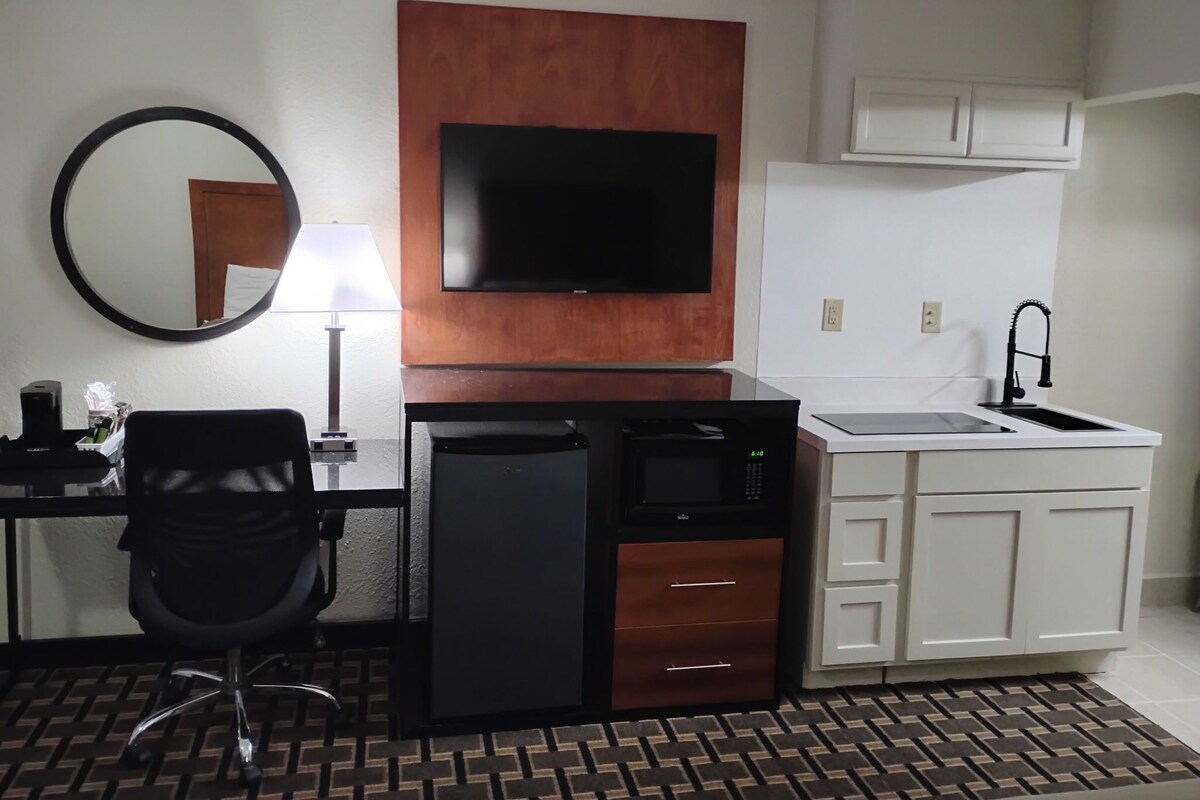 A compact hotel room with a kitchenette, a flat-screen TV mounted on a wooden panel, a round mirror, a desk with a chair, and a small sink.