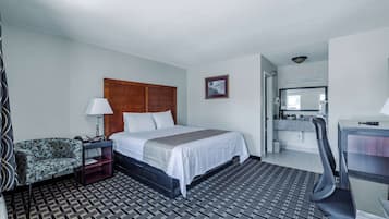 Quarto de hotel com cama, escrivaninha, cadeira, mesa de cabeceira, abajur, tapete estampado e um quadro na parede.