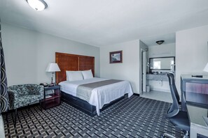 Een hotelkamer met een bed, een bureau, een stoel, een nachtkastje, een lamp, een kamerkleed met een patroon en een ingelijste foto aan de muur.