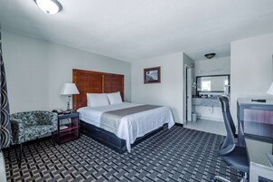 Habitación de hotel con cama, escritorio, silla, mesita de noche, lámpara, alfombra estampada y un cuadro en la pared.