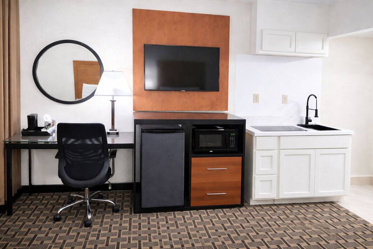 A compact hotel room with a desk, chair, flat-screen TV, mini-fridge, microwave, and a small kitchenette with a sink.