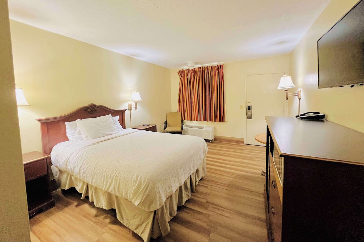 A hotel room with a large bed, bedside tables, a desk, a chair, a TV, and a door with curtains.