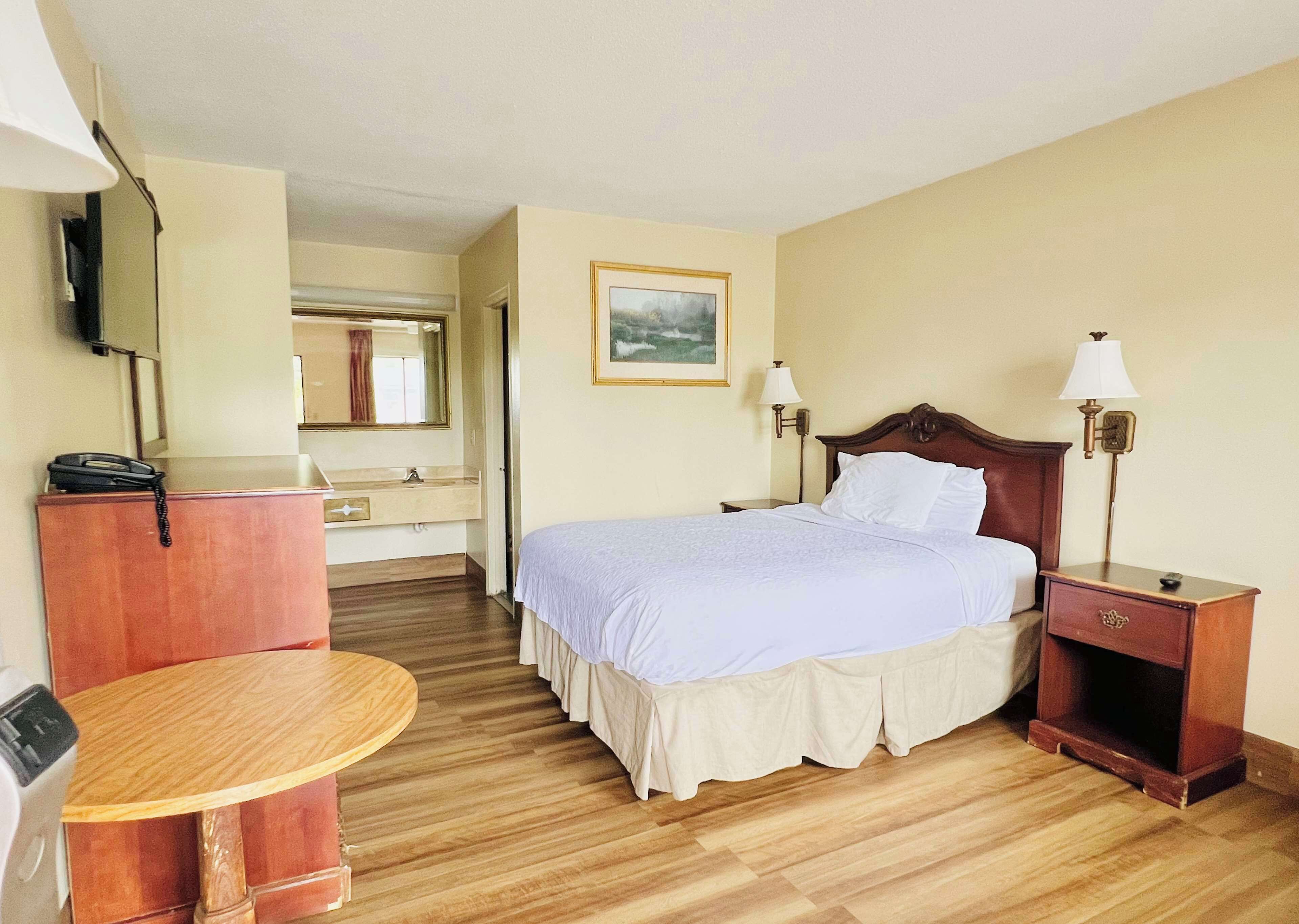 A hotel room with a bed, bedside lamps, a wooden nightstand, a round wooden table, a mirror, and a painting on the wall.
