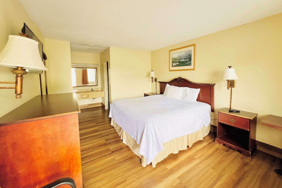 A hotel room with a wooden floor, a bed with white bedding, a wooden dresser, a nightstand, and a lamp.