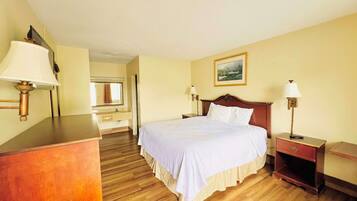 A hotel room with a wooden floor, a bed with white bedding, a wooden dresser, a nightstand, and a lamp.