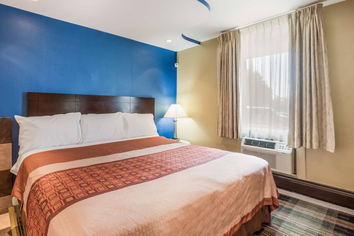 A hotel room with a large bed, a nightstand, a lamp, an air conditioning unit, and a window with curtains.