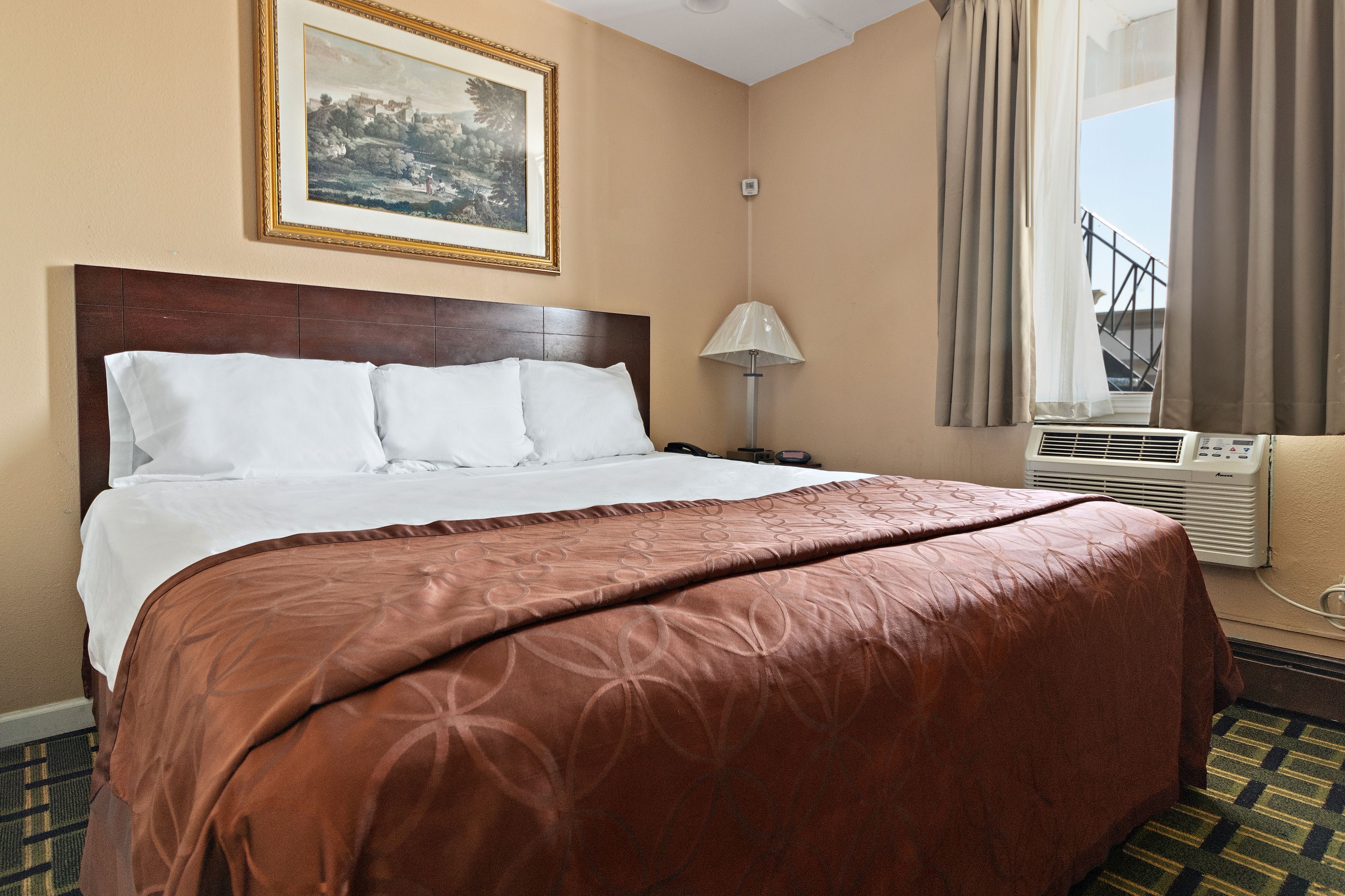 A neatly made bed with a brown bedspread, a framed picture on the wall, a lamp, and an air conditioning unit.