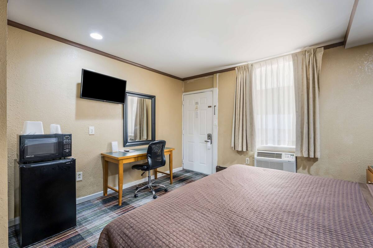 A hotel room with a bed, a desk with a chair, a microwave, a television, and an air conditioning unit.