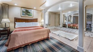 A hotel room with a large bed, a separate bathroom with a bathtub, and a dining area visible through an archway.