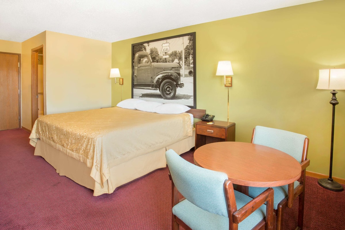 A hotel room with a bed, a round table with chairs, a desk, two lamps, and a wall-mounted picture of a vintage car.