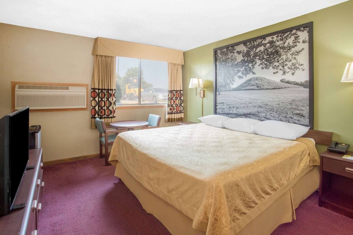 A hotel room with a large bed, a TV, a desk with a chair, a window with curtains, and a painting on the wall.