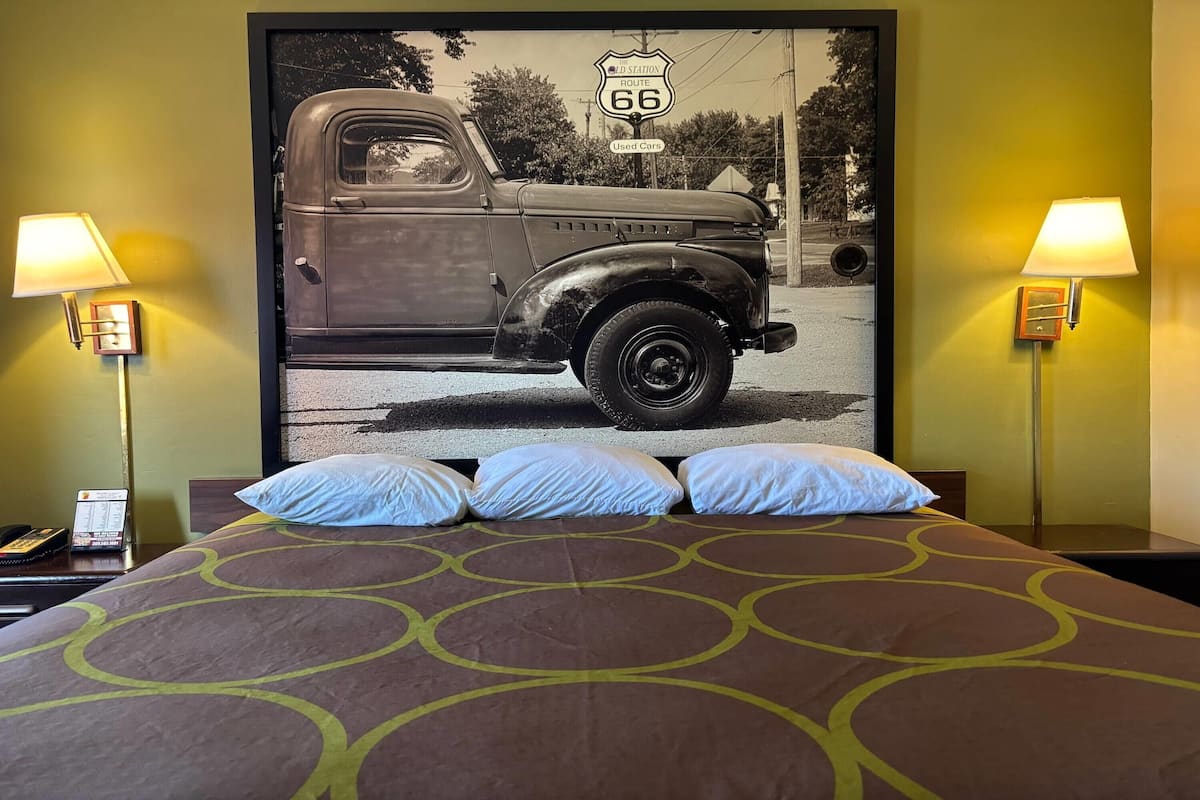 A hotel room with a bed, two bedside lamps, a telephone, and a large vintage car poster on the wall.